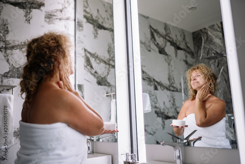 Foto A senior woman with curly hair stands in a stylish bathroom, applying moisturizing cream to her neck while looking at her reflection in the mirror