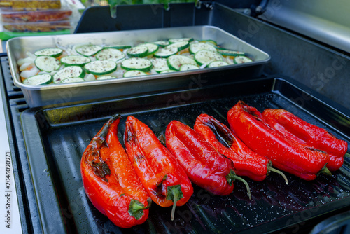 Fresh red pointed peppers and seasoned zucchini slices are cooking on a barbecue grill. A close-up of a gas grill loaded with fresh vegetables.