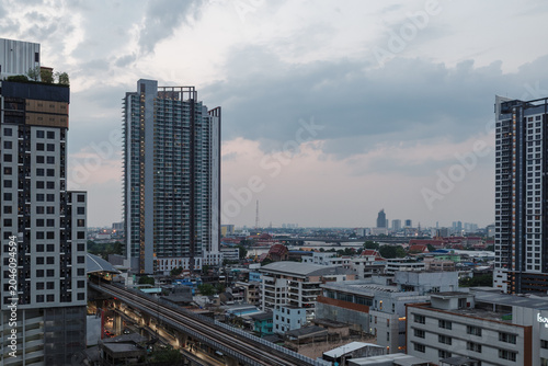 A high-angle, wide shot of an urban landscape in Bangkok, Thailand, featuring prominent modern high-rise residential towers, an elevated transit railway line cutting through the city, and a sprawling 