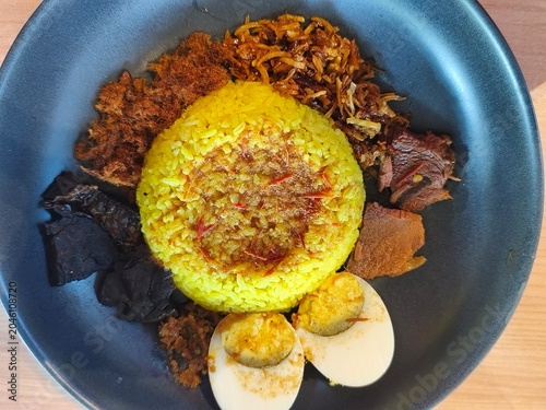 Nasi Kuning, Yellow rice with additional pieces of boiled egg, chili beef, fermented soybean cake, fried money, and chili sauce on a black plate on a wooden table 