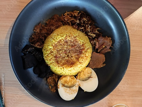 Nasi Kuning, Yellow rice with additional pieces of boiled egg, chili beef, fermented soybean cake, fried money, and chili sauce on a black plate on a wooden table 