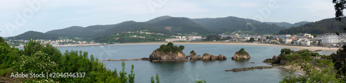 Foto Os Castelos rock formations on Covas Beach in Viveiro, Galicia, showcasing mossy coastal stones, seagulls on the sand, and a hillside town in the background