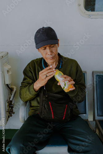 A passanger asian man in a black hat and sling bag sitting relax in a bench opening a fresh bottle orange water drink.