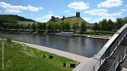 A serene summer day in Vilnius, Lithuania, showcasing the historic Gediminas' Tower overlooking the calm Neris River, with a lone jogger enjoying the peaceful riverside path.