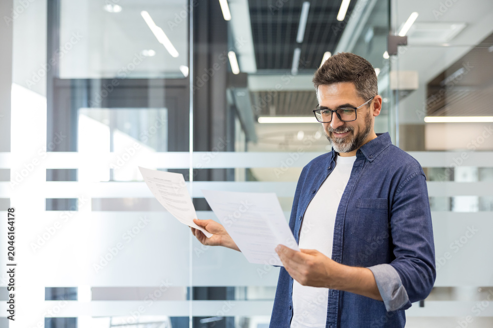 Fototapeta premium Confident man reviewing corporate paperwork in a contemporary office setting, focusing on data analysis, planning, and achieving business goals with a positive expression