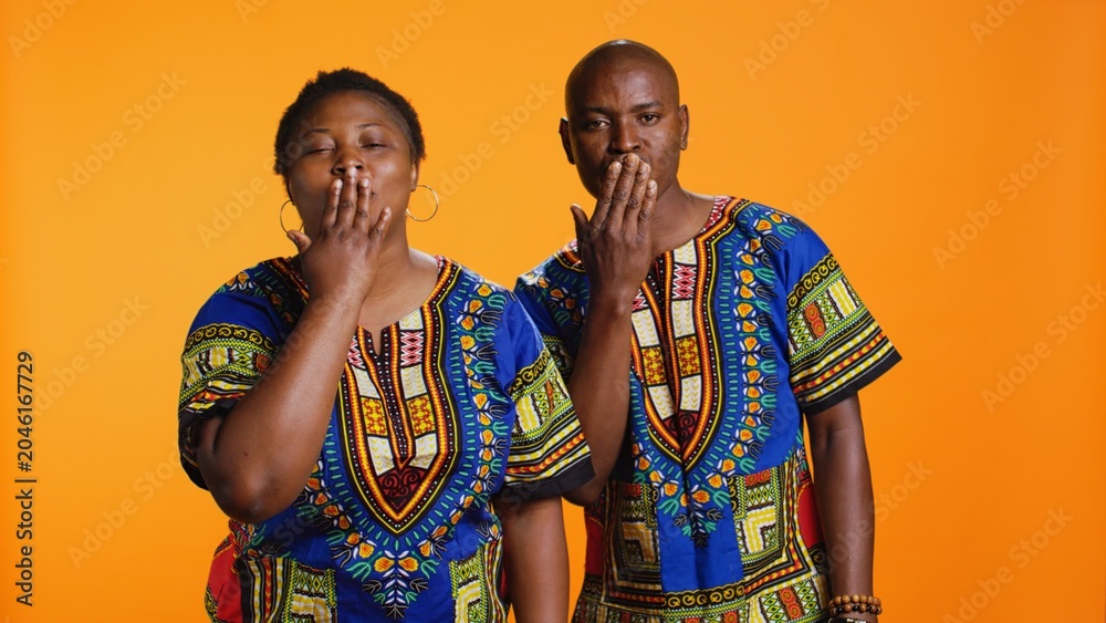 Obraz premium Sweet african american couple sending air kisses on camera, presenting flirty gesture and showing their affection. Ethnic man and woman blowing kiss in studio, traditional clothing items.