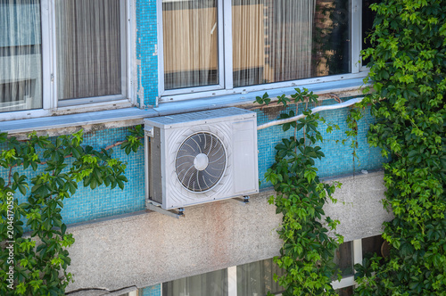 The air conditioner's outdoor unit is on the exterior of the apartment building. Ivy plants covering the exterior of the building.
