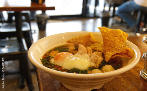 Thai spicy Tom Yum noodle soup with soft boiled Onsen egg served in a white bowl at a restaurant table.