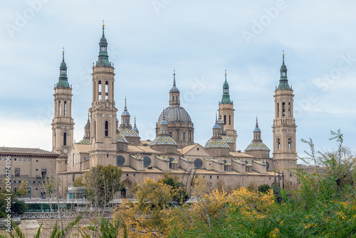 Majestic Basilica of Our Lady of the Pillar framed by green trees and autumn foliage in Zaragoza, Spain. The historic Spanish cathedral features stunning domes and towers under a soft overcast sky