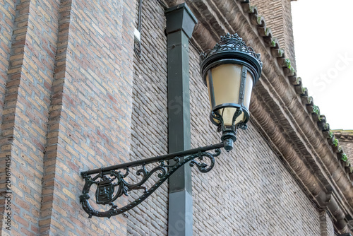 Vintage ornate street lamp attached to a classic brick wall in the historic center of Zaragoza, Spain. Detailed view of traditional Spanish wrought iron lighting fixture