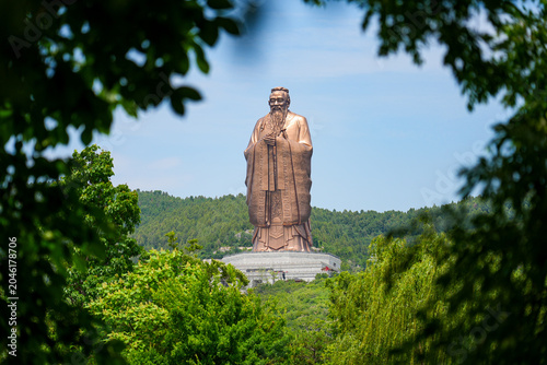 Фотография Giant statue of Confucius at Nishan Shengjing framed by green tree leaves, Qufu,