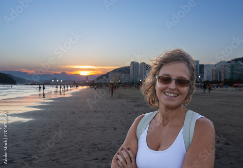 Smiling woman enjoying sunset at Santos Beach, Brazil, with urban skyline in the background. Travel, leisure, coastal lifestyle and tourism concept.