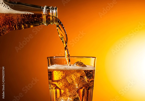 Amber liquid pouring from bottle into glass filled with ice cubes, studio lighting