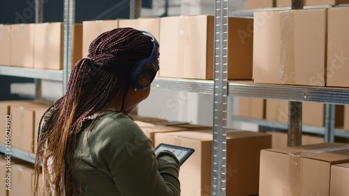 Slika na platnu Chilled african american warehouse laborer listening music while checking labels on cardboard boxes