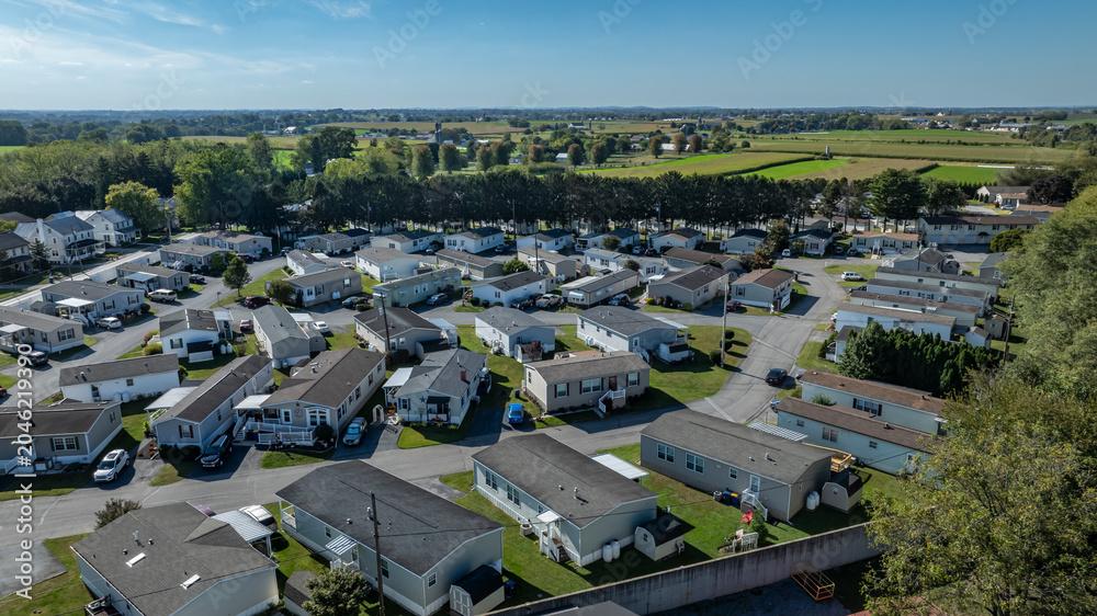 Obraz premium People live in a mobile and manufactured home community with rows of houses in a green rural area. The sun shines on the neighborhood and fields surround the homes. Cars are parked along the streets.