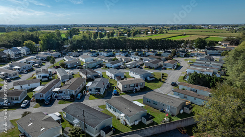 People live in a mobile and manufactured home community with rows of houses in a green rural area. The sun shines on the neighborhood and fields surround the homes. Cars are parked along the streets.