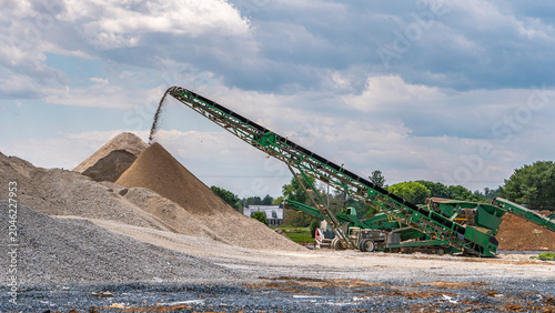 A machine lifts and moves crunched concrete and sand at a construction site. Workers are present around the piles of materials. The sky is cloudy, and trees are visible in the background.