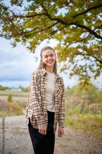 Smiling young woman standing on rural path under autumn tree, casual lifestyle portrait with natural background, warm colors and peaceful countryside atmosphere