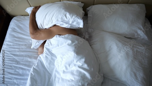Fotografija Young man sleeping in large bed and covering head with pillow in modern hotel room or home interior