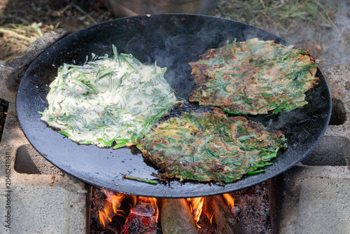 Traditional Korean chive pancakes cooking on a large iron griddle over a wood fire outdoors with smoke rising and rustic countryside cooking atmosphere.