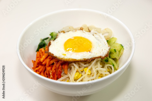 Traditional Korean bibimbap topped with fried egg, bean sprouts, vegetables, and seasoned side ingredients served in a white bowl on a clean background.