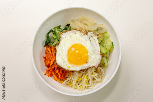 Traditional Korean bibimbap topped with fried egg, bean sprouts, vegetables, and seasoned side ingredients served in a white bowl on a clean background.