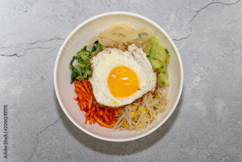 Traditional Korean bibimbap topped with fried egg, bean sprouts, vegetables, and seasoned side ingredients served in a white bowl on a clean background.