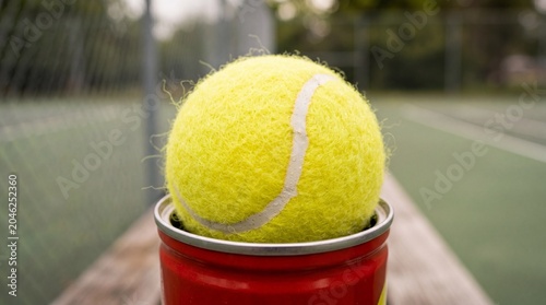 A fresh yellow tennis ball sits atop a red metal can against a blurred green court. It conveys a sense of preparation, fresh beginnings, and eagerness for athletic practice.