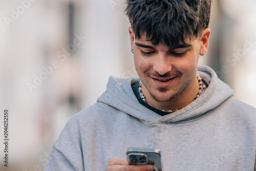 Young hispanic man smiling while using his smartphone outdoors, wearing a casual urban outfit, urban background, informal lifestyle