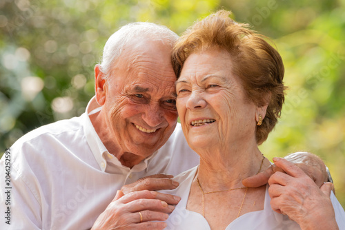 Joyful elderly caucasian couple smiling and embracing in a sunny outdoor setting, radiating love, happiness, and togetherness in their golden years