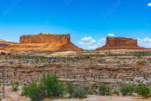 Fotografie Two flat-topped red sandstone buttes tower over sparse desert scrubland and layered rock flats near the Canyonlands National Park entrance, Utah