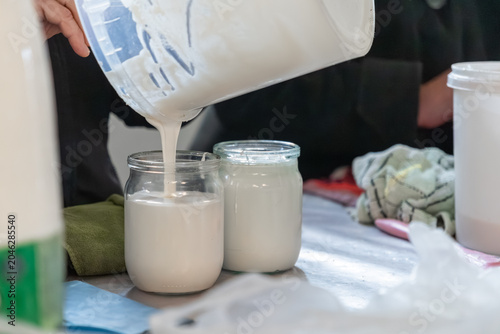A person pours fresh creamy sour cream into a glass jar from a plastic bucket, showcasing the careful preparation of organic homemade dairy products for sustainable kitchen storage
