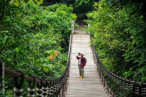 A Solo Hiker Uses Binoculars While Standing On A Wooden Suspension Bridge In A Lush Tropical Rainforest During A Wilderness Adventure