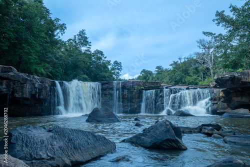 Wide Waterfall Cascading Over Rocky Ledges Into A Calm Forest Stream During A Sunny Day
