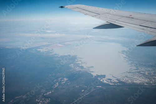 Tableau sur toile Aerial view shows pale salt pan with dark scrubland and faint road lines in Botswana, airplane wing at upper right, daylight haze softens distant horizon
