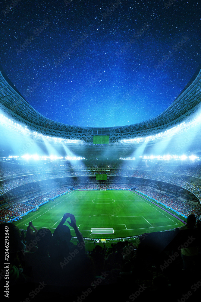 Obraz premium Modern massive modern soccer stadium at night, brightly lit by floodlights. Cheering crowds fill the tiers under a deep blue starry cosmos. Silhouetted fans are in the foreground. Football match