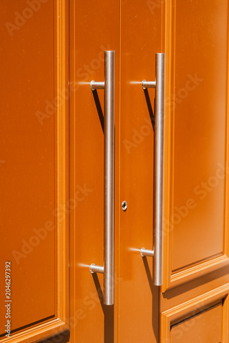 Close-up of a painted orange double door featuring two vertical stainless steel pull handles mounted on raised panels. Warm directional lighting casts soft