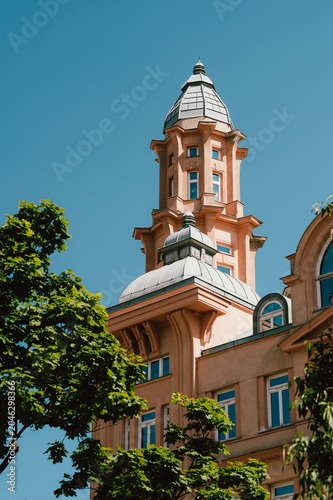 A pale peach historic tower with ornate turrets and a domed roof rises above a tree-lined city street against a clear blue sky. Decorative cornices, arched