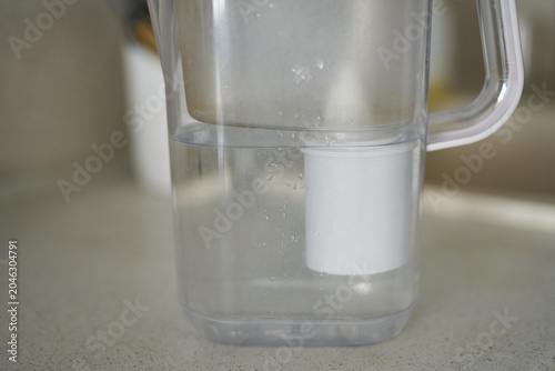 Filtered Water Pitcher Centered On Kitchen Countertop Showing White Filter Cartridge Submerged In Clear Water Inside Plastic Jug Near Faucet In Neutral Domestic Kitchen Scene.