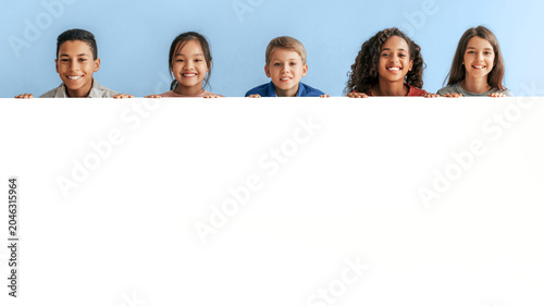 Canvastavla Happy Multicultural Group Of Five Children, Including Boys And Girls, Smiling And Peeking Over A White Banner, In A Bright Studio Setting