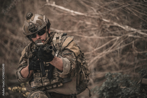 Armed tactical operator aiming rifle during military combat mission in outdoor nature environment. Professional private military contractor conducting security patrol operation in forest wilderness.