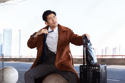 Fotografija Exhausted businessman traveler sitting with suitcase and loosening tie in city