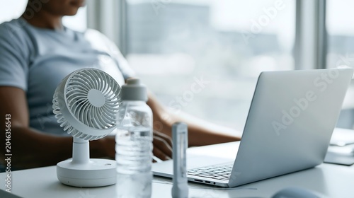 Office worker using portable fan beside laptop and water bottle during hot weather indoors. Concept of workplace heat management, hydration and employee wellbeing