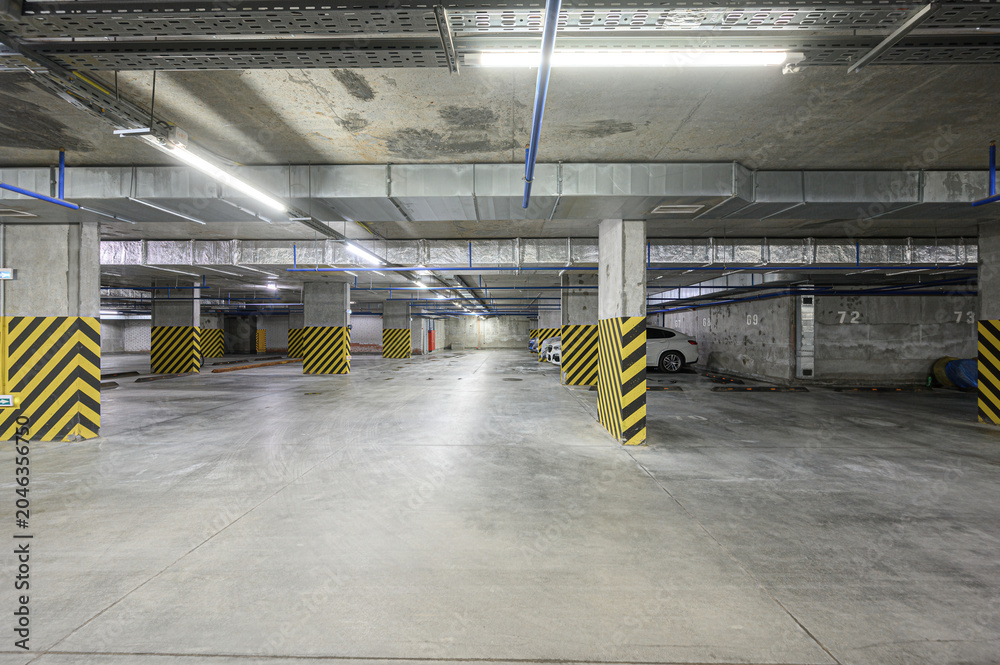 Naklejka premium Spacious underground parking with concrete pillars wrapped in yellow-black hazard stripes, blue overhead pipes, fluorescent lighting, and a few parked cars in marked bays