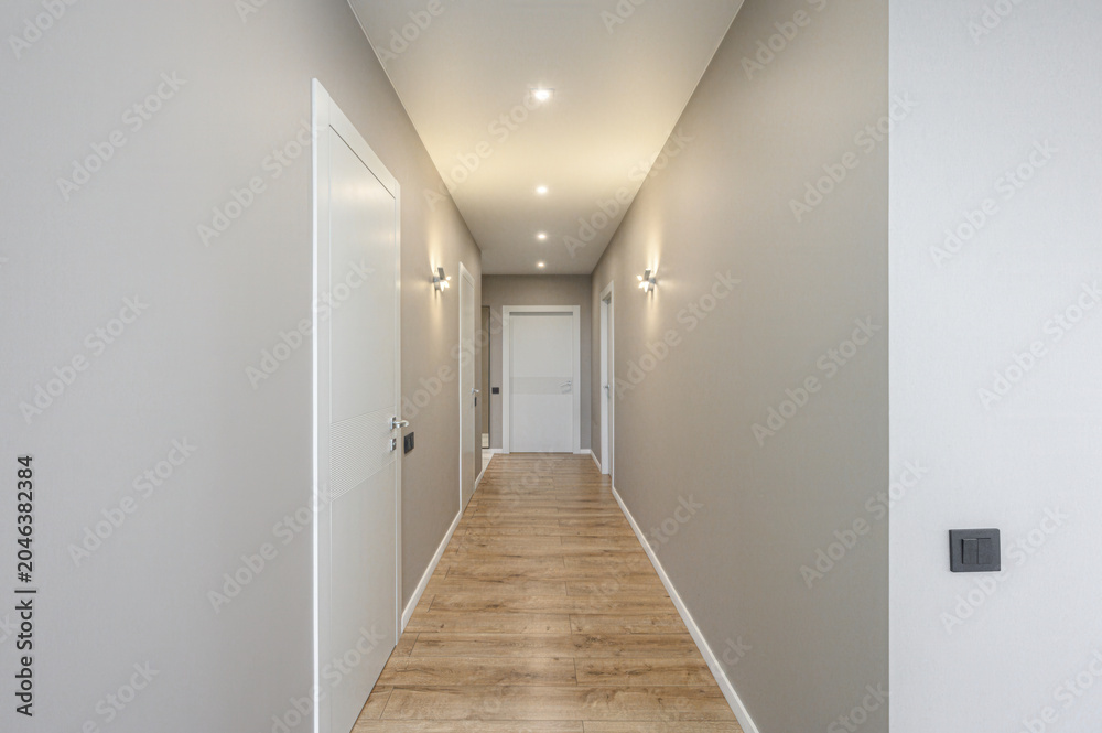 Fototapeta premium narrow modern hallway with light grey walls, warm wooden laminate flooring, white interior doors, recessed ceiling lights, and stylish wall sconces; minimalist design