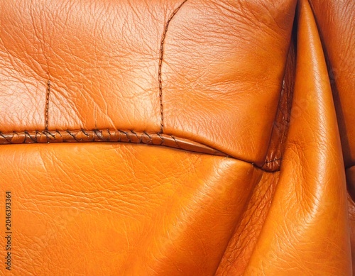 Close-up of richly textured, vibrant orange-brown leather. Detailed stitching accents folds and seams, creating a tactile and warm aesthetic