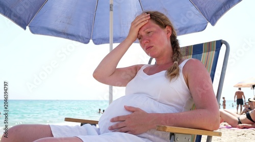 Pregnant woman sitting under a beach umbrella and holding her forehead while resting outdoors. Pregnancy health, heat exhaustion, summer wellness and maternal care concept.