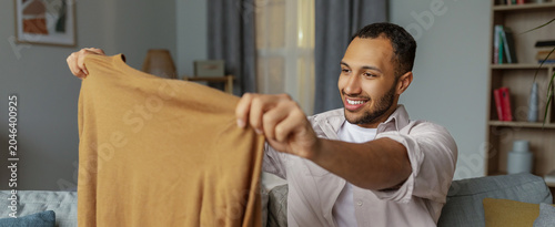 Bild auf Leinwand Smiling Young Man Of Mixed Race Holding Sweater While Sitting In Cozy Living Room, Enjoying Home Comforts