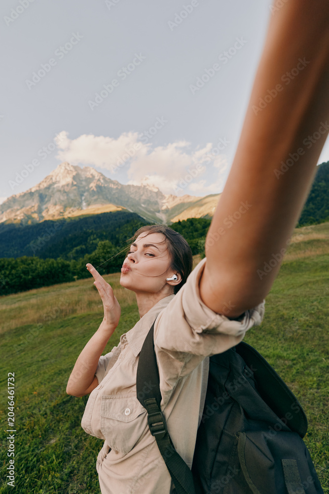 Naklejka premium Young woman outdoors raises her arm for a selfie, smiling with eyes closed as the sun lights a grassy hillside with distant mountains, wearing a beige shirt and backpack.