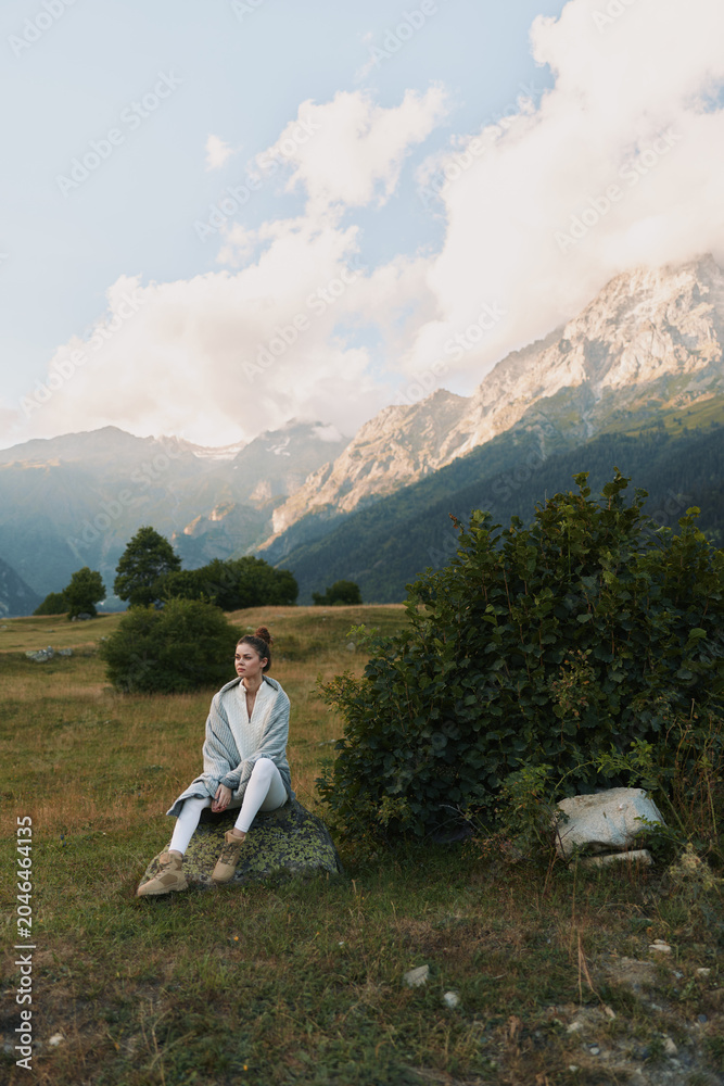 Naklejka premium Woman lifestyle sitting on rock in mountain meadow, casual outfit, thoughtful expression, natural light, scenic landscape, cloudy sky, peaceful atmosphere